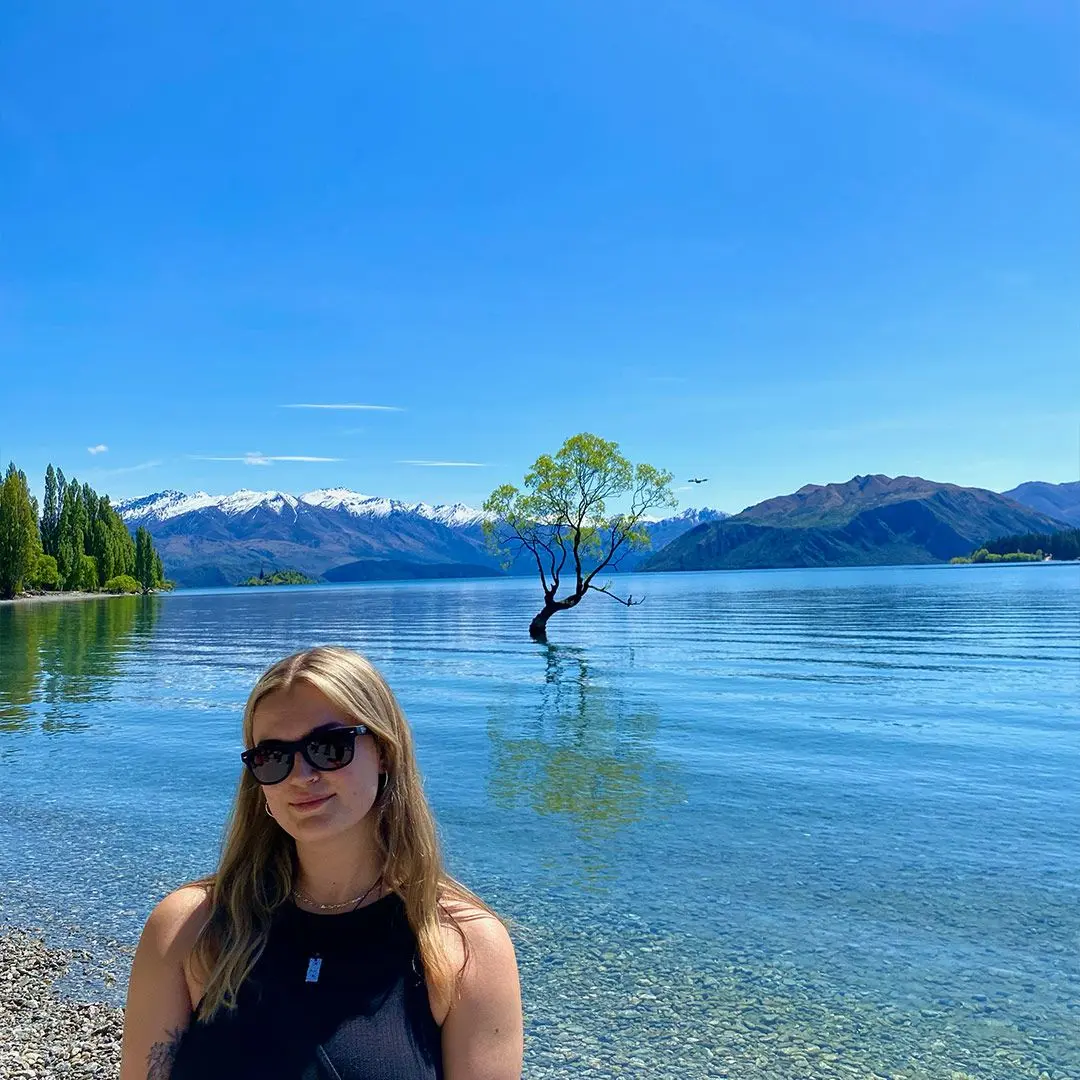 Blonde girl in front of lake with blue sky and tree in background.
