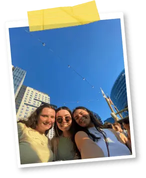 Three girls (one wearing sunglasses) taking a selfie under a blue sky and tall buildings.png
