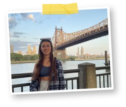 Girl in an open checkered shirt standing beside railing with river and bridge in the background, clue sky